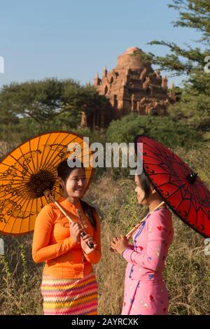 Un modèle de tournage avec deux jeunes femmes en robe traditionnelle et parasols avec un petit temple en arrière-plan à Bagan, au Myanmar. Banque D'Images