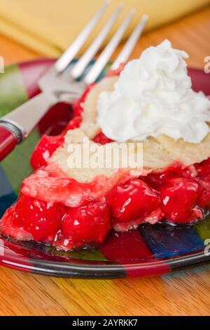 Tarte aux cerises avec crème fouettée, sur une plaque de verre rayée avec une fourchette rouge et une serviette jaune sur une table en bois Banque D'Images