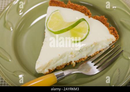 Tranche de tarte à la chaux, avec une tranche de citron vert torsadé sur le dessus, reposant sur une plaque de chine sur une nappe à motif fleuri Banque D'Images