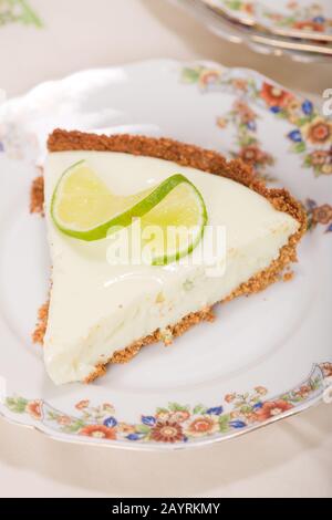 Tranche de tarte à la chaux, avec une tranche de citron vert torsadé sur le dessus, reposant sur une plaque de chine sur une nappe à motif fleuri Banque D'Images