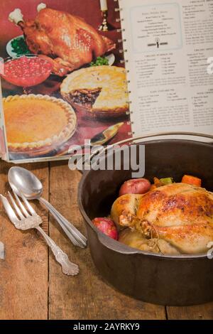 Poulet rôti de style néerlandais avec pommes de terre rouges cuites, carottes et céleri, dans une casserole en fonte sur une table en bois rustique, avec un vieux livre de cuisine dans le dos Banque D'Images