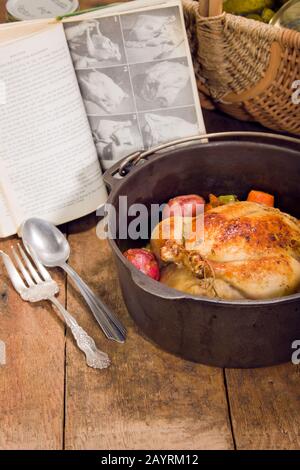 Poulet rôti de style néerlandais avec pommes de terre rouges cuites, carottes et céleri, dans une casserole en fonte sur une table en bois rustique, avec un ancien livre de cuisine, un panier et c Banque D'Images