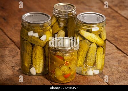 Trois bocaux de cornichons d'aneth et un pot de cornichons doux, montrant les concombres marinés, les mauvaises herbes, l'ail, les poivrons rouges et les feuilles de raisin, sur un W rustique Banque D'Images