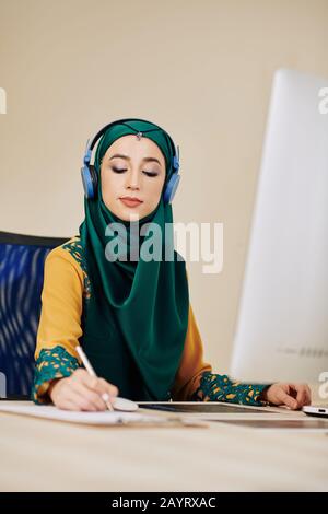 Entrepreneur féminin en hijab écoute de la musique dans les écouteurs lors de travail sur le projet Banque D'Images