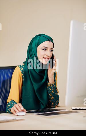 Portrait du jeune ingénieur logiciel musulman parlant au téléphone avec le client et en vérifiant les détails du projet Banque D'Images