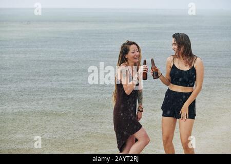 De jeunes femmes heureuses dansant sous la pluie sur la plage et des bouteilles de bière de clinking Banque D'Images