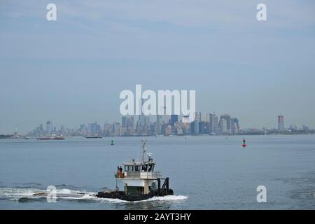 Un petit bateau « baignoire » utilitaire qui tracte un traîneau à eau sur Upper Bay, vu de la vue classique de la ligne d'horizon de Manhattan sur Staten Island Ferry, en toile de fond Banque D'Images