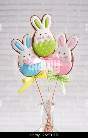 Groupe de trois biscuits de lapin de pâques sur bâton en bois dans vase en verre avec mur de briques blanches derrière. Pain d'épices maison recouvert de glaçage coloré pour la décoration de table. Banque D'Images