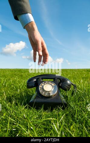 Main d'un homme d'affaires inconnu atteignant pour le récepteur d'un téléphone rotatif noir à l'ancienne assis dans une herbe verte luxuriante Banque D'Images