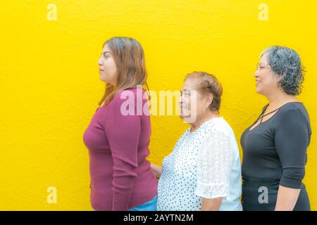Profils de vue latérale de la grand-fille et de la fille, trois générations de femmes mexicaines souriant avec des vêtements décontractés dans une rangée sur un dos jaune Banque D'Images