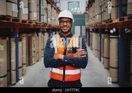 Un travailleur souriant en portrait portant un gilet orange et un casque blanc avec des bras croisés dans un grand entrepôt souriant Banque D'Images