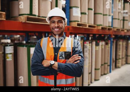 Homme africain confiant debout avec des bras repliés dans l'unité de fabrication avec casque blanc et uniforme en usine Banque D'Images