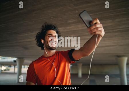Jeune homme gai debout sous le pont en faisant un appel vidéo sur son smartphone avec écouteurs - jeune homme prenant un selfie tout en faisant de l'exercice Banque D'Images