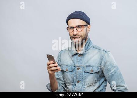 Âge moyen attrayant modèle masculin vêtu de poils avec des vêtements à la mode, est titulaire d'smart phone, conversations avec des collègues ou amis en réseaux socail Banque D'Images