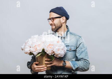 De horizontales heureux homme barbu regarde ailleurs pendant que détient bouquet de fleurs blanches, d'être dans la bonne humeur comme a ce jour avec sa petite amie, rend présent Banque D'Images