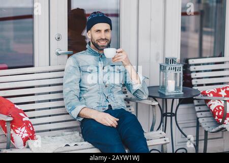 Un homme aux yeux bleus heureux avec un look positif, se trouve face à un intérieur confortable à la cafétéria de la terrasse, aime le café aromatique, a plus de temps après le travail. Beau Banque D'Images