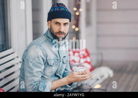 Homme barbu d'âge moyen avec yeux bleus, barbe épaisse et moustache, porte un chapeau élégant et une veste denim, tient une tasse de thé chaud ou de café, a réfléchi Banque D'Images