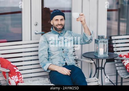 Photo d'un homme barbu aux yeux bleus, vêtu de vêtements en denim, lève la main avec une tasse de café ou de thé, de bonne humeur, repose dans un cafeter extérieur Banque D'Images