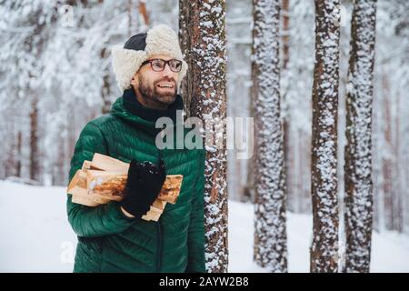 Agréable à heureux homme détient le bois de chauffage, l'air songeur de côté, se dresse près de arbres d'hiver, des rêves à propos de quelque chose d'agréable, jouit de magnifiques wint Banque D'Images