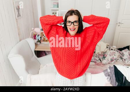 Portrait horizontal de cheerful brunette femme au foyer porte des lunettes et pull chaud rouge, pose à l'intérieur d'accueil dans sa chambre, a sourire positif. Banque D'Images