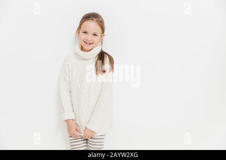 Portrait de heureux satisfait girl porte chandail tricoté blanc chaud, a l'expression joyeuse, se dresse contre l'arrière-plan studio avec copie vierge de l'espace. Adora Banque D'Images