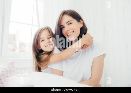 Tourné à l'intérieur de la bonne à la mère brunette avec sourire doux et sa petite fille donne aux hug, profitez de l'atmosphère intérieure, pose contre blanc spacieux Banque D'Images