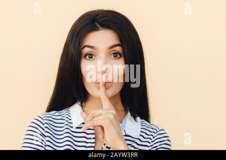 Secret femme fait le signe de la rousse, des commissionns avec le meilleur ami, garde index doigt sur les lèvres, a de longs cheveux sombres, pose sur fond de studio. Femme mignonne t Banque D'Images