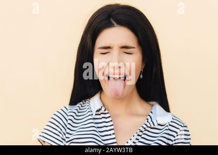 Portrait de belle femme fait grimace, maintient la bouche large ouverte et montre la langue, garde les yeux fermés, porte des vêtements décontractés, pose contre le studio bac Banque D'Images