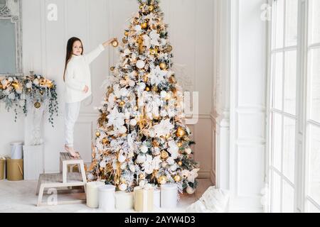 Une photo intérieure d'adorable petit enfant se tient sur la chaise sur les pieds à dos, décorera le grand arbre du nouvel an, a de bonne humeur, étant en prévision des vacances et des désirs Banque D'Images