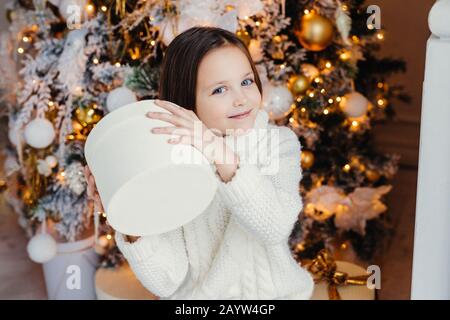 Blue Eyed joli joli petit enfant tient présent fort, se demande ce qu'est à l'intérieur, se dresse près de l'arbre de Noël ou Nouvel An, reçoit des parents. surprise Banque D'Images