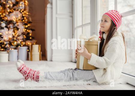 Portrait de femme reposant sur le côté enfant porte chandail blanc, un pantalon et des chaussettes chaudes, englobe des cadeaux emballés, est assis sur le plancher en chambre douillette, admire Nouveau Banque D'Images