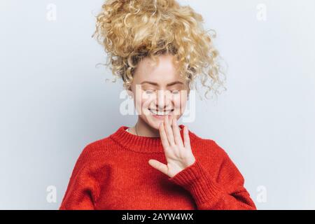 Portrait de heureuse jeune femme enchantée avec une expression gaie, garde les yeux fermés et sourire agréable, vêtu d'un pull rouge, lève la main et le geste Banque D'Images