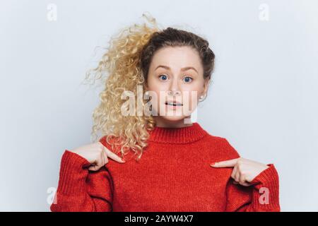 Jolie jeune femme dans le bewilderment porte rouge chaud ample chandail, indique l'espace vierge de copie de vêtements pour votre contenu publicitaire, a poney t Banque D'Images