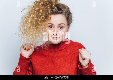 Photo horizontale de belle femelle drôles à yeux bleus avec des cheveux raboueux et bouclés dans la queue de poney, maintient les mains enfoncées dans les poings, les élève, a inattendu Banque D'Images