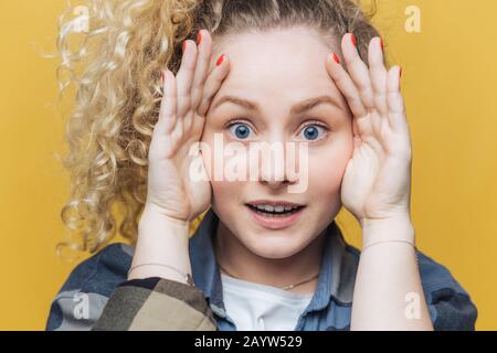 Gros plan sur une jolie femme émotionnelle avec une peau saine et des yeux bleus, garde les mains sur la tête, regarde scrupuleusement et étonnamment à l'appareil photo, les remarque ainsi Banque D'Images