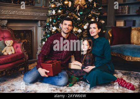 Tourné à l'intérieur de la famille les jeunes ont une bonne relation, s'asseoir ensemble sur le plancher près de Nouvel An des arbres décorés, tenir présent, féliciter les uns les autres avec com Banque D'Images