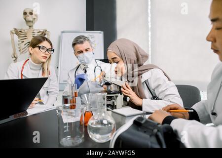 Groupe de scientifiques médecins chimistes faisant des enquêtes, travaillant en laboratoire. Jeune chercheuse de femme musulmane regardant au microscope. Ses collègues Banque D'Images