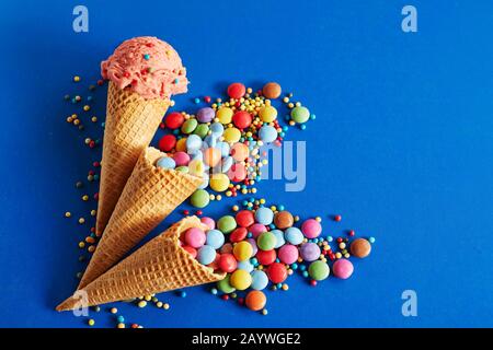 Assortiment de bonbons colorés débordant de cônes de crème glacée avec une seule pelle de crème glacée aux fraises sur un fond bleu avec un espace de copie pour un festi Banque D'Images