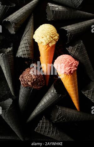 Crème glacée aux fraises, au chocolat et à la vanille dans des cônes de gaufre, sur fond noir et blanc de cornets gris dans une couche plate Banque D'Images