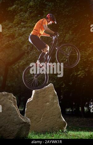 Spectacle extrême de cycliste talentueux qui se produit sur de grandes roches debout sur la roue arrière, en soirée dans le parc, vue latérale, concept de mode de vie actif Banque D'Images