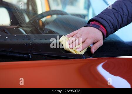 L'homme après le lavage essuie le pare-brise de sa voiture orange avec un chiffon jaune au lavage de voiture. Main mâle et carrosserie de voiture près. Banque D'Images