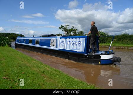 Narrowboat appelé Ivar III en passant le long du canal Shropshire Union à Cheshire sous un soleil lumineux et sous le ciel bleu avec un homme au timon. Banque D'Images