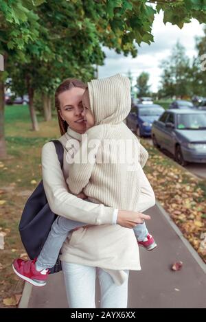 la jeune famille, la femme, la mère porte l'enfant dans ses bras, le petit garçon dans le sweat avec une capuche, la ville de marche, le concept d'élever un soin d'enfant et l'amour Banque D'Images