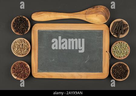 Six petits bols en bois remplis de divers types de grains de poivre, une cuillère de cuisson en bois faite de bois d'olive et un vieux plateau en ardoise noire avec copie de spac Banque D'Images