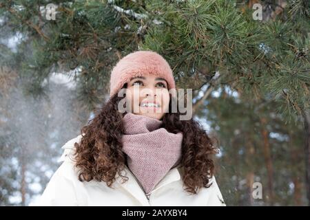 fille caucasienne aux cheveux bouclés bruns portant une élégante tenue d'hiver debout sous des branches de pins souriant, portrait horizontal à bas angle Banque D'Images