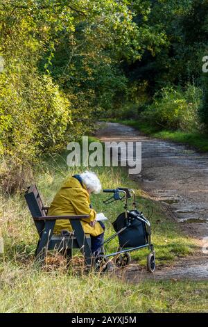 Une vieille dame âgée avec un zimmer ou un cadre de marche assis sur un banc en bois lisant un livre parmi les arbres dans certains bois par elle-même. Banque D'Images