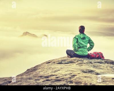 Belle femme de corps assis dans un windcheater vert sur le sommet de montagne regardant le lever du soleil sur une mer de brouillard Banque D'Images