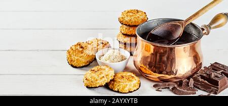 Cookies à la noix de coco trempés dans du chocolat fondu dans une casserole de cuivre avec barre de chocolat hachée et noix de coco déshydratée dans un bol avec du bois blanc en wi Banque D'Images