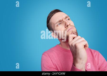 Jeune homme contemplatif barbu touchant le menton et regardant vers le haut contre fond bleu Banque D'Images