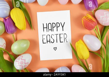 Carte texte « Happy Easter » entourée d'œufs de pâques colorés et de fleurs de printemps tulipes sur fond orange, plat Banque D'Images